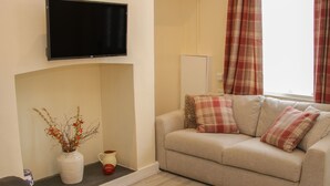A living room with a sofa, a flat-screen TV mounted on the wall, a fireplace, and a window with curtains. - Whitewater Cottage (Llangollen)