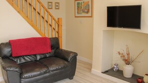 A living room with a black leather sofa, a red throw blanket, a wooden staircase, a flat-screen TV mounted on the wall, and a vase with branches on a shelf. - Whitewater Cottage (Llangollen)