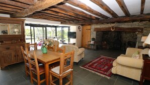 A rustic living room with exposed wooden beams, a stone fireplace, a dining area with wooden furniture, and large windows offering a view of the outdoors. - Garth Morthin The Farmhouse (Porthmadog)
