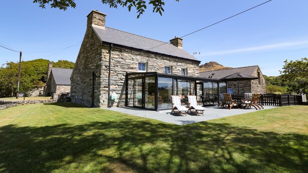 A stone house with a glass conservatory, outdoor seating, and a stone chimney. - Garth Morthin The Farmhouse (Porthmadog)