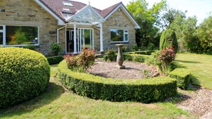 A stone house with a conservatory, surrounded by well-manicured hedges and a garden.