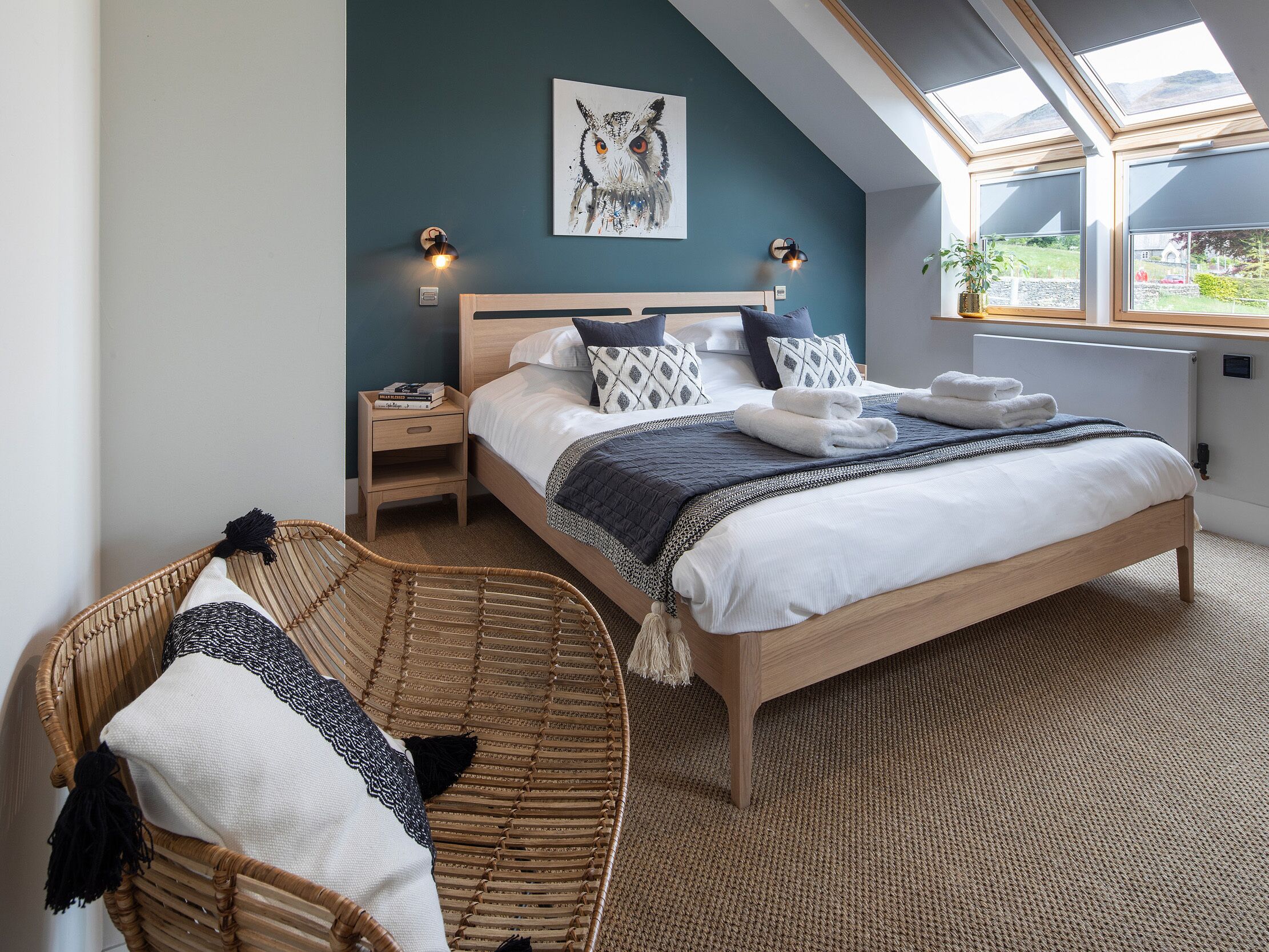 A bedroom with a bed, bedside tables, a wall-mounted lamp, a window with a view, and a woven chair.
