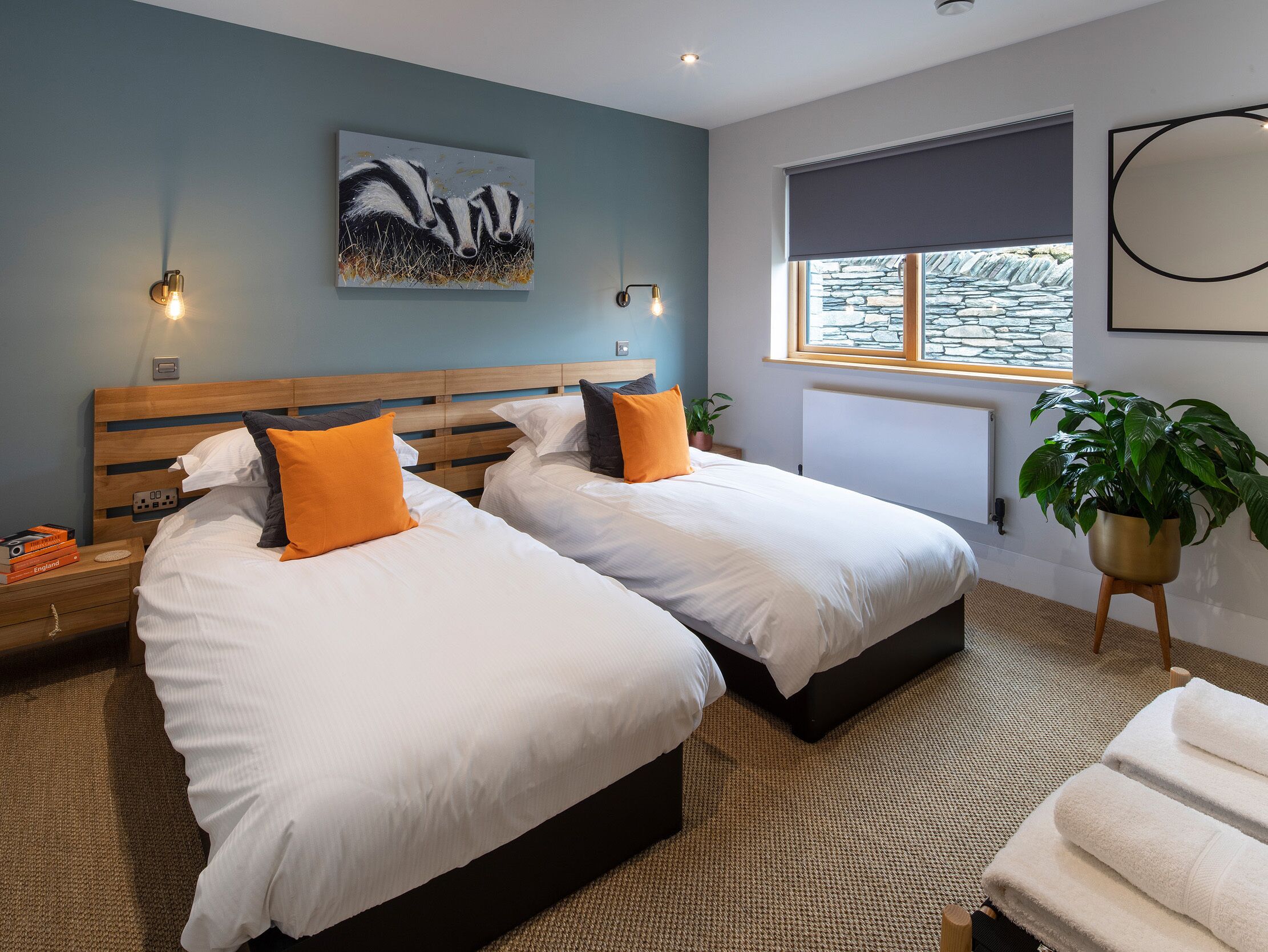 A hotel room with two beds, a painting of badgers, a potted plant, and a window with a view of stone walls.