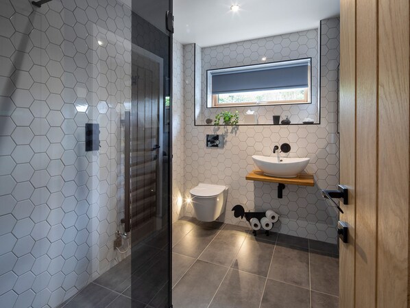 Une salle de bain moderne avec des carreaux hexagonaux, une douche en verre, des toilettes, un lavabo avec une étagère en bois et un porte-rouleau de papier toilette en forme de chien.