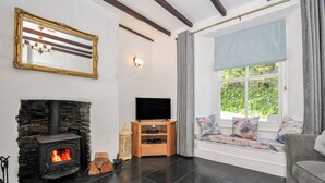 A living room with a stone fireplace, a television on a wooden stand, and a window with a view of greenery. - Fairfield Cottage (Boscastle)