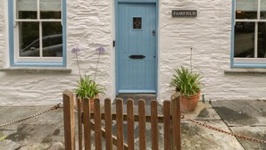 A white building with a blue door and two windows, a sign reading "FAIRFIELD", and two potted plants in front. - Fairfield Cottage (Boscastle)