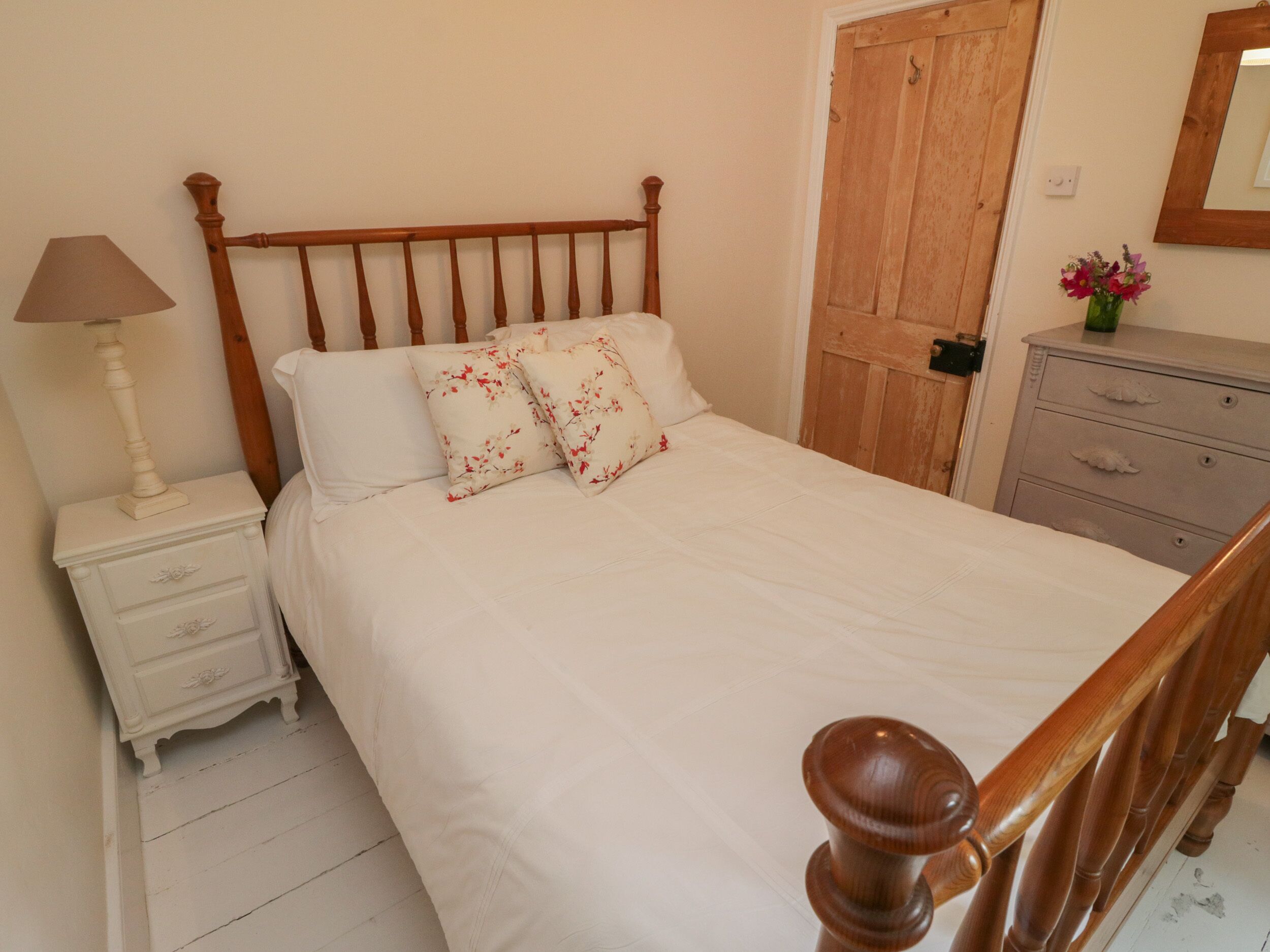 A bedroom with a wooden bed frame, white bedding, a nightstand with a lamp, a dresser, and a wooden door.