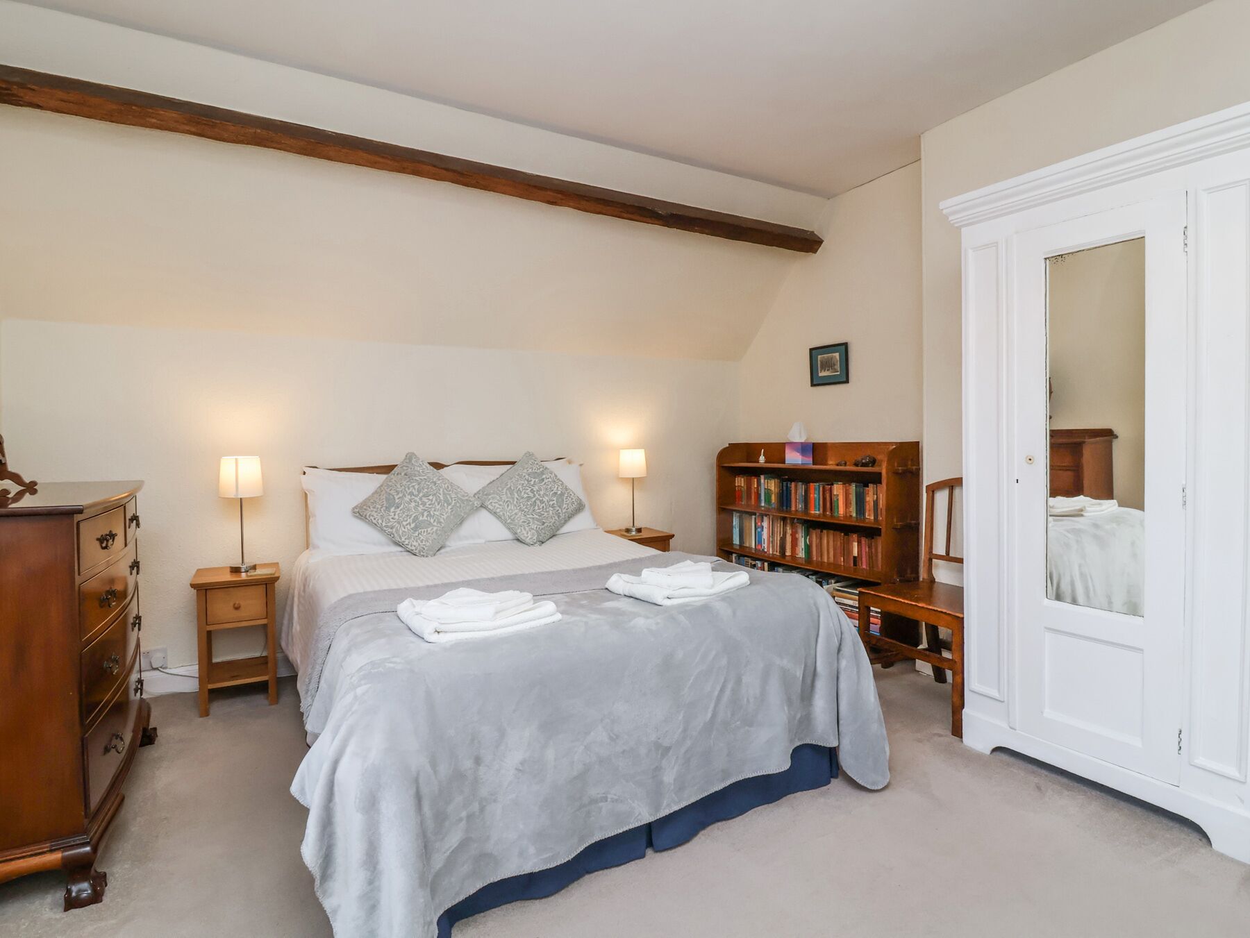 A bedroom with a bed, two bedside tables, a wooden dresser, a bookshelf, and a white door with a mirror.