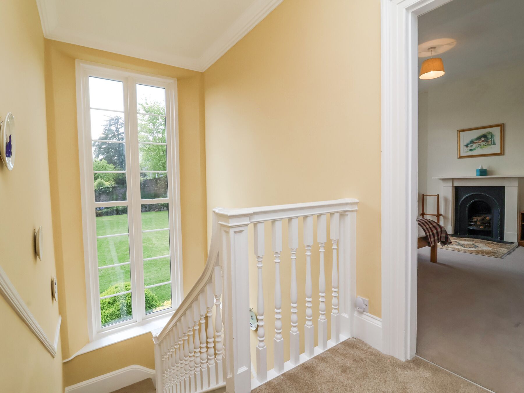 A staircase with white balusters leading to a landing. A fireplace with a mantel and a framed picture above it is visible in the adjacent room.