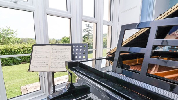 A grand piano with a music stand and sheet music in front of a large window overlooking a garden. - The Old Vicarage (Tiverton)