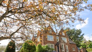 A large, two-story brick house with a symmetrical facade, multiple windows, and a well-maintained garden with various shrubs and a lawn. - The Old Vicarage (Tiverton)