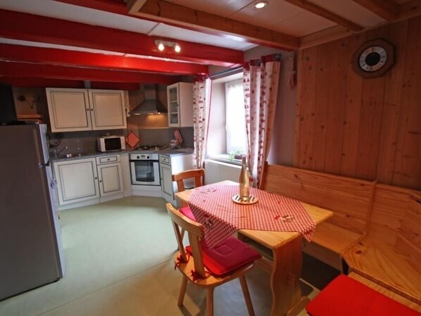 Cocina compacta con armarios blancos, microondas y estufa. Hay una mesa de madera con mantel a cuadros y una silla con cojín rojo.