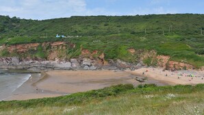 Uma praia costeira com falésias rochosas, uma área de areia e uma colina verde ao fundo.