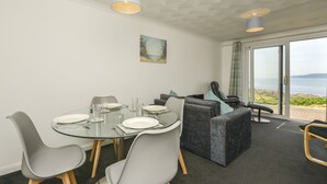 Uma sala de jantar com uma mesa de vidro, cadeiras cinzentas e um sofá. Há um quadro na parede e vista para o mar pela porta de vidro.