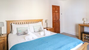 Ein ordentlich bezogenes Bett mit blauer Decke, Holz-Kopfteil und dekorativen Kissen. Ein Nachttisch mit Lampe, eine Holzbank und eine Tür sind im Hintergrund zu sehen.
