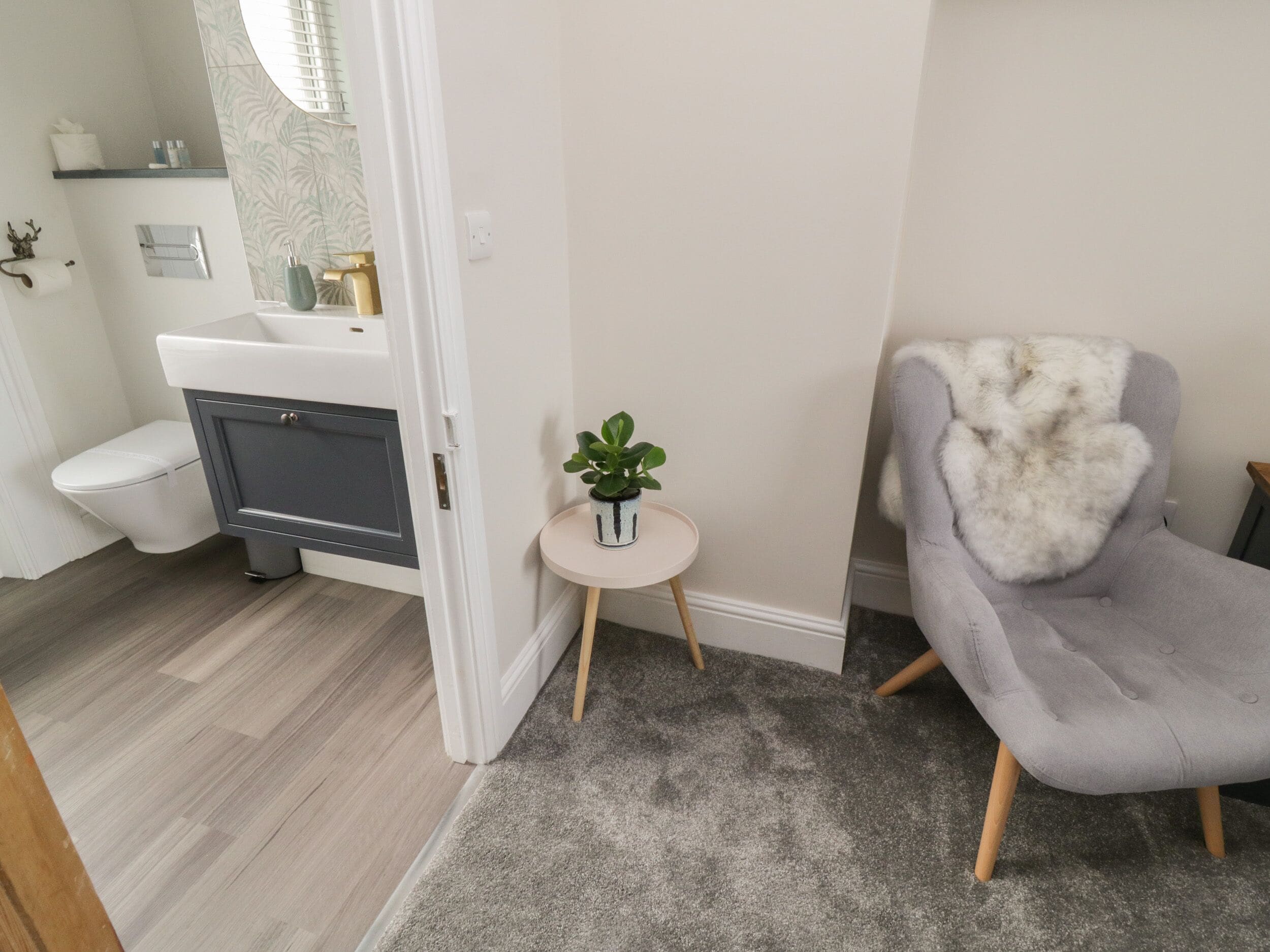 Een badkamer met toilet, wastafel en douche. Er staat een grijze fauteuil en een klein rond tafeltje met een plant.