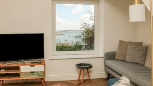 A living room with a TV, a small wooden side table, a sofa, and a window with a view of boats and hills.