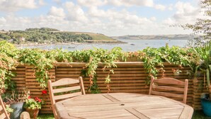 A wooden table and chairs set on a balcony overlooking a harbor with boats and hills in the distance.