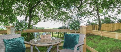 An outdoor wooden deck with wicker furniture and a table, surrounded by greenery and a wooden fence.