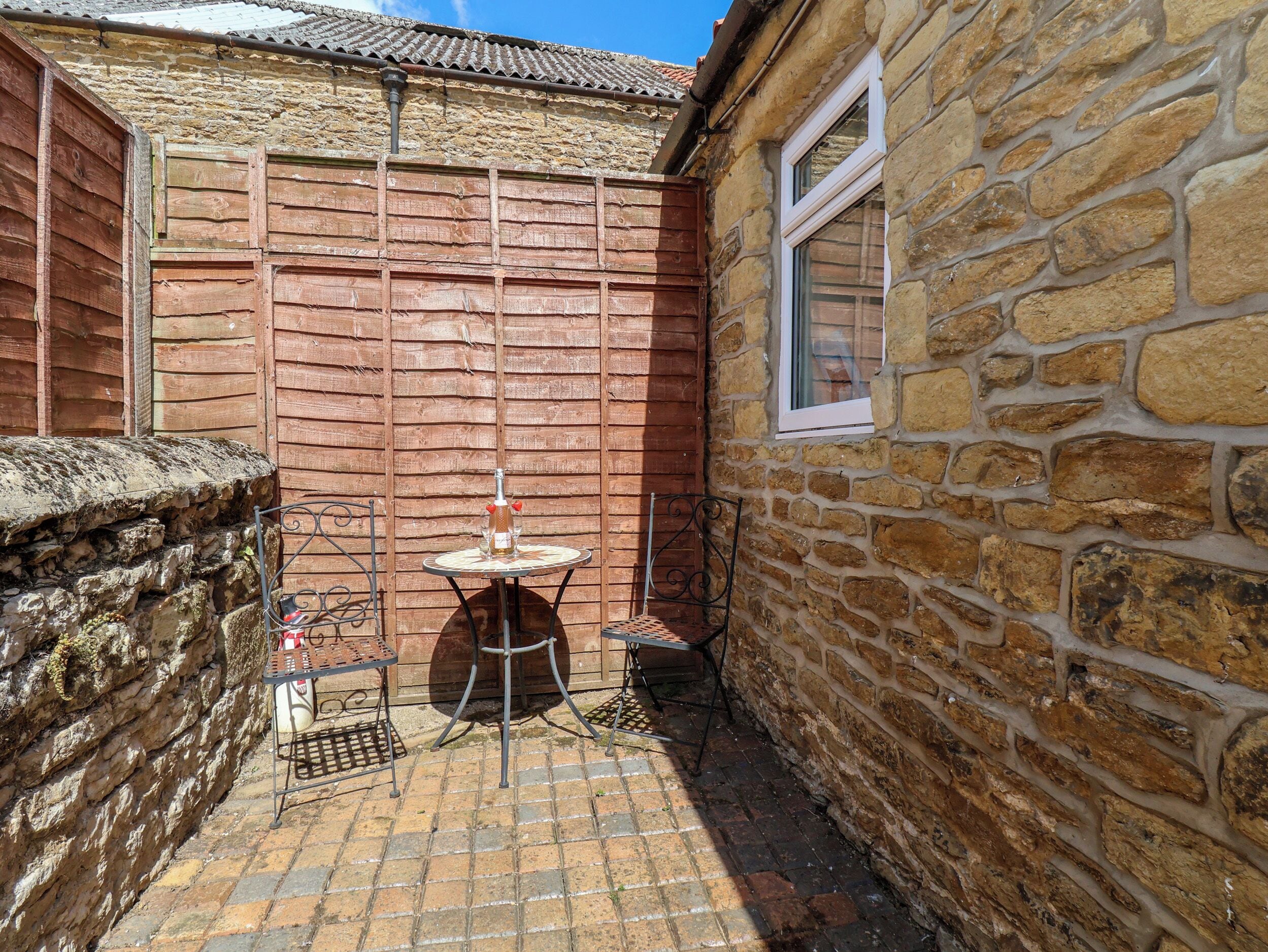 A stone-walled courtyard with a small round table and chairs, a bottle, and a glass.