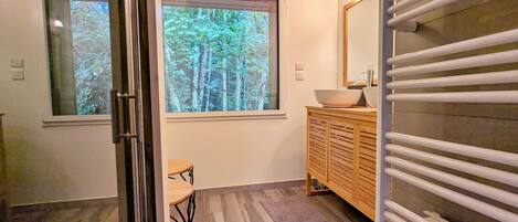 A modern bathroom with a wooden vanity, a large window with a forest view, and a glass door leading to another room.