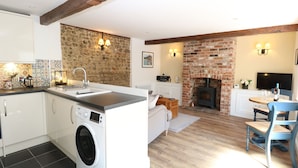 A modern kitchen with a built-in washing machine, a stone wall, and a brick fireplace. - Captain's Cottage (Sheringham)