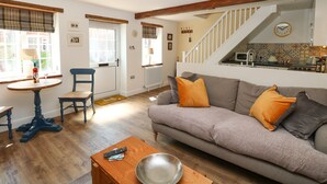 A cozy living room with a sofa, a coffee table, and a dining area with a table and chairs. A staircase leads to an upper level. - Captain's Cottage (Sheringham)