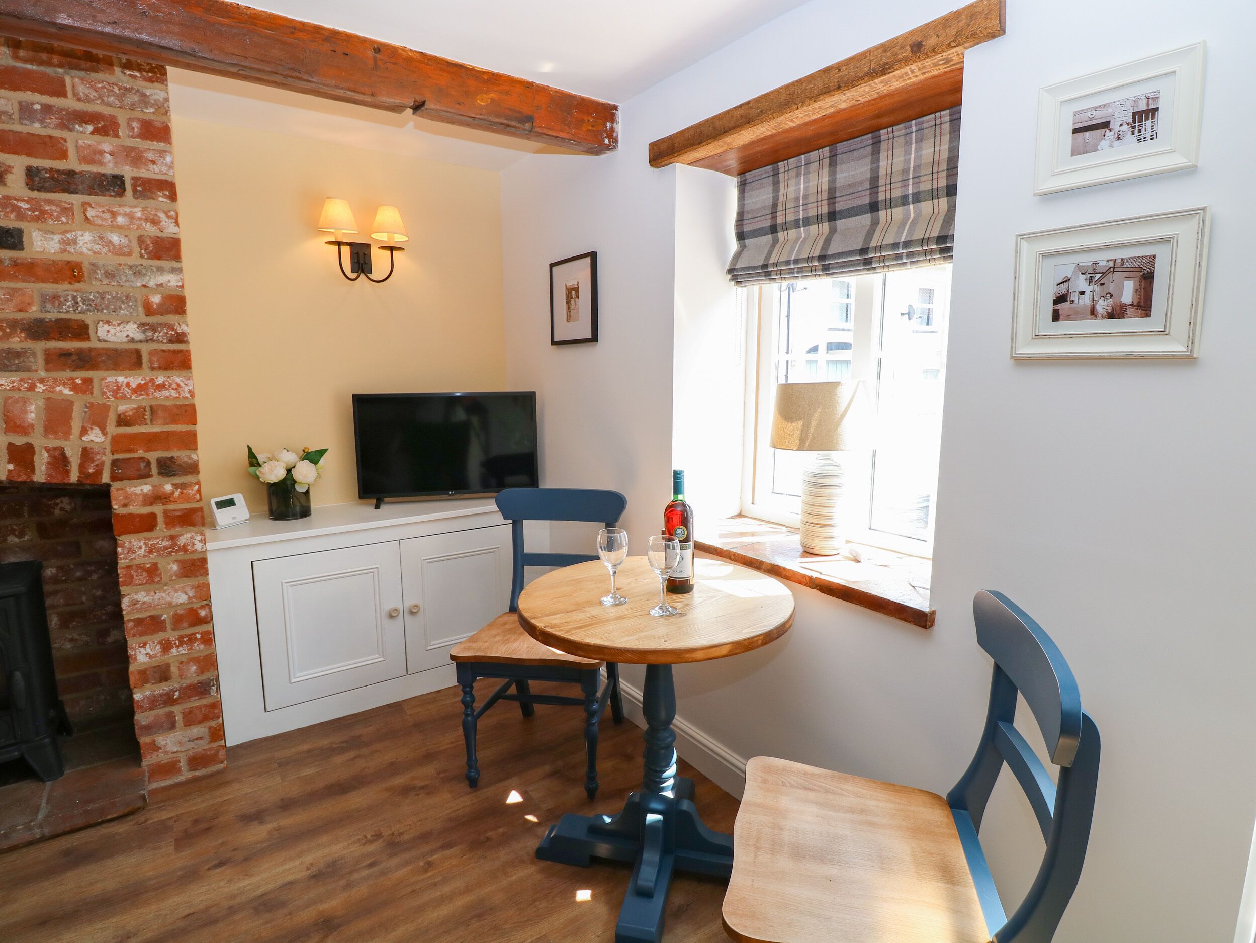 A cozy room with a brick fireplace, a television, a round wooden table with chairs, and a window with a plaid blind.