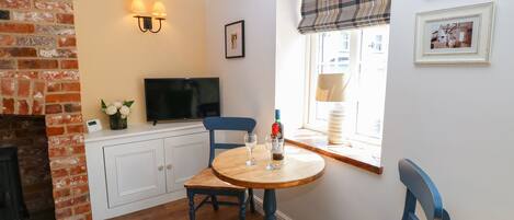 A cozy room with a brick fireplace, a television, a round wooden table with chairs, and a window with a plaid blind.