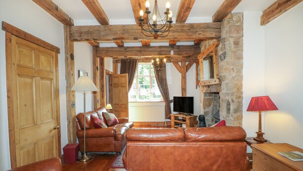 A rustic living room with wooden beams, leather sofas, a stone fireplace, and a television. - Lees Moor Cottage (Matlock)