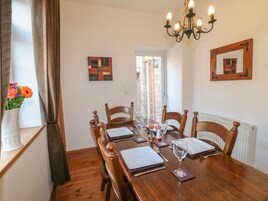 Uma sala de jantar com uma mesa de madeira posta para quatro pessoas, rodeada por cadeiras. Há um vaso com flores num parapeito, um candeeiro e obras de arte nas paredes.