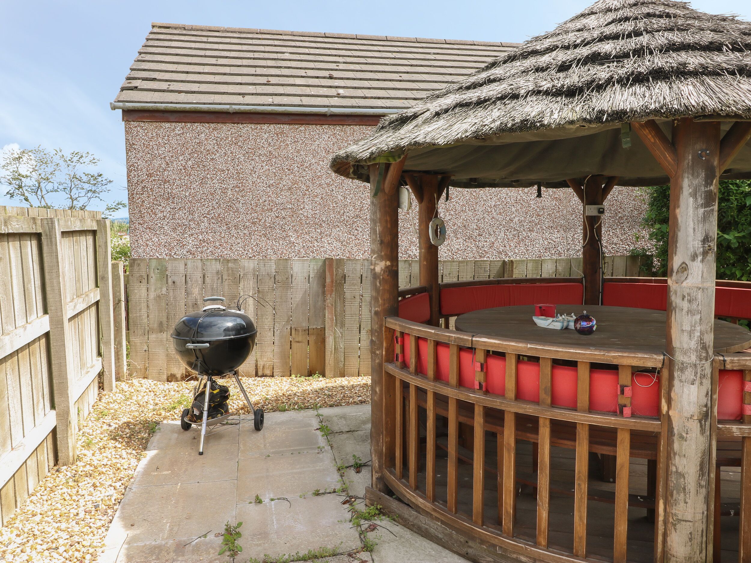 A wooden gazebo with a thatched roof, a barbecue grill, and a red cushioned seating area.