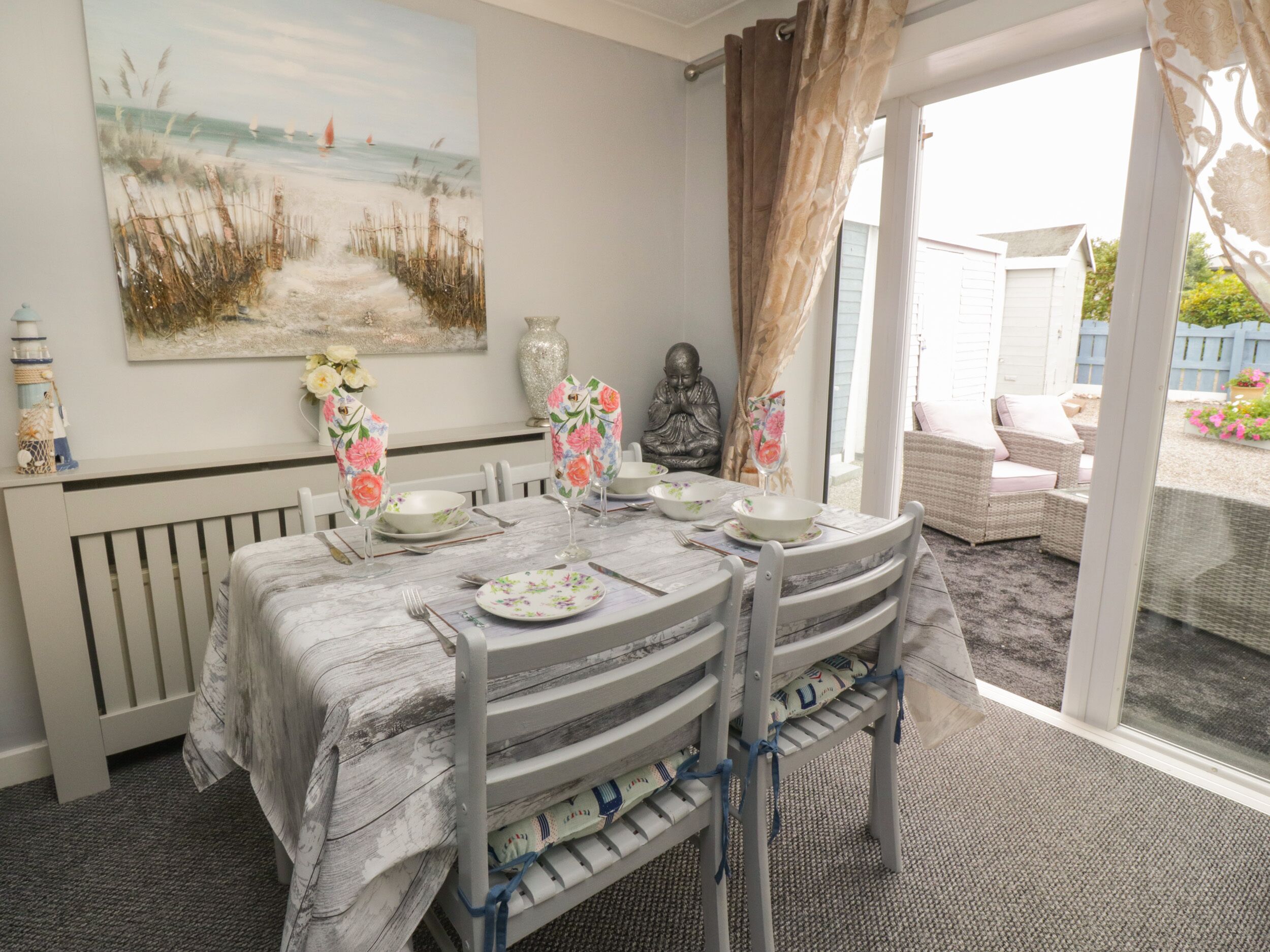A dining area with a table set for four, a beach-themed painting on the wall, and a view of the garden through the glass door.