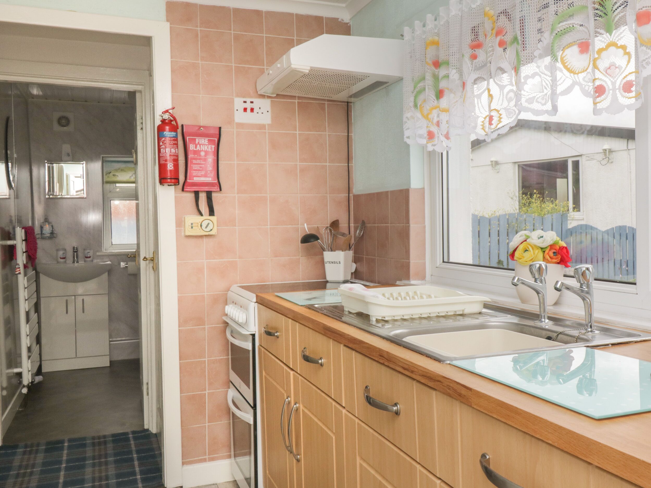 A kitchen with a wooden countertop, a sink, and a window with lace curtains. There is a fire blanket and a fire extinguisher on the wall.
