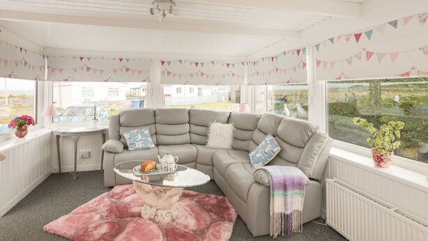 A cozy living room with a grey sectional sofa, a glass coffee table, and a pink rug. There is a window with a view of a garden and a white radiator. - Sandpiper (Dumfries)