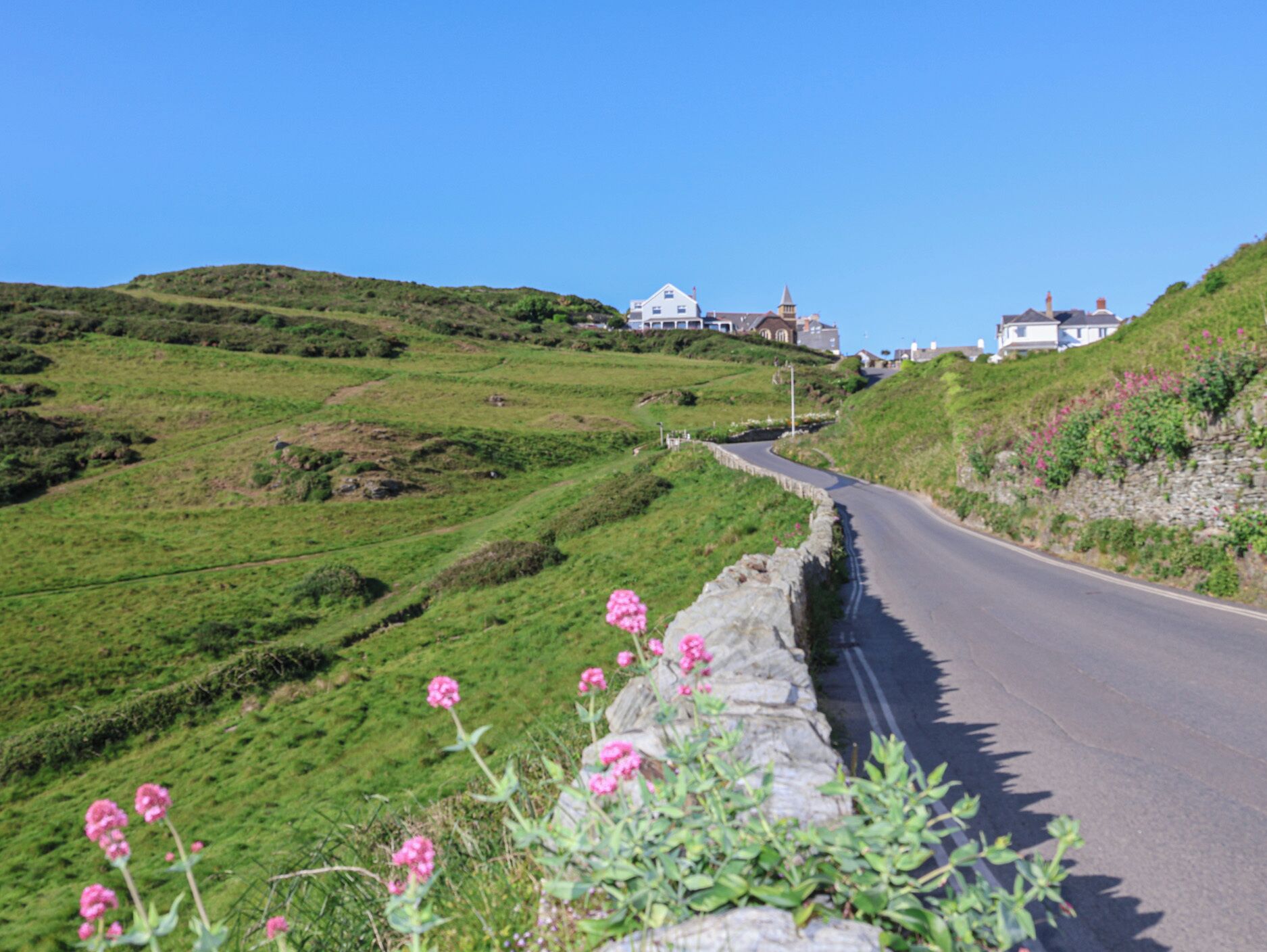 A rural road curving through green hills with stone walls and houses in the distance.