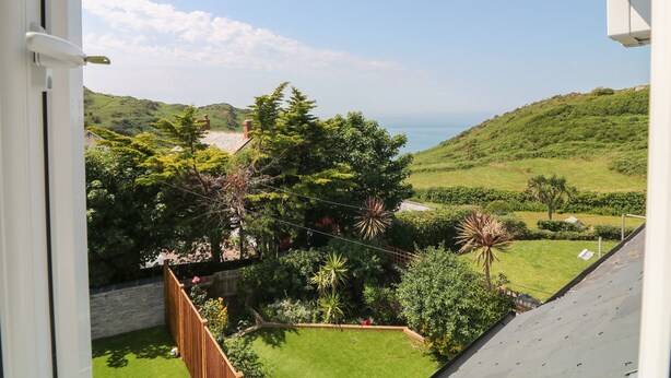 Vista da janela sobre um jardim exuberante, com diversas árvores e arbustos, um gramado bem cuidado e uma vista distante do mar.
