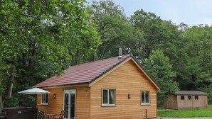 Una pequeña cabaña de madera con camino de grava, un patio con mobiliario exterior y zonas boscosas alrededor.