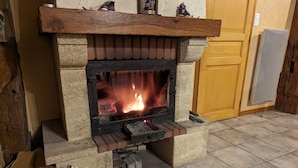 A stone fireplace with a fire burning inside, a wooden mantel above it, and a tiled floor. - Charming Nature Retreat: Cozy Cottage with Fireplace and Scenic Views (Girmont-Val-d'Ajol)