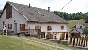 A two-story house with a sloped roof, a wooden fence, and a picnic table in the front yard. - Charming Nature Retreat: Cozy Cottage with Fireplace and Scenic Views (Girmont-Val-d'Ajol)