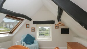 A bedroom with a sloped ceiling, a bed with a floral bedspread, a chair with a blue cushion, a wooden dresser, and a window with a view of houses. - Athelstan Cottage (Stroud)