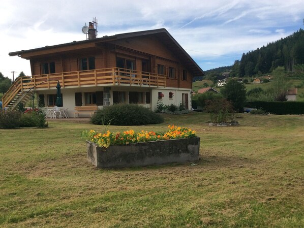 Una casa de madera de dos pisos con un balcón, rodeada por un jardín cubierto de césped y una cama de flores.