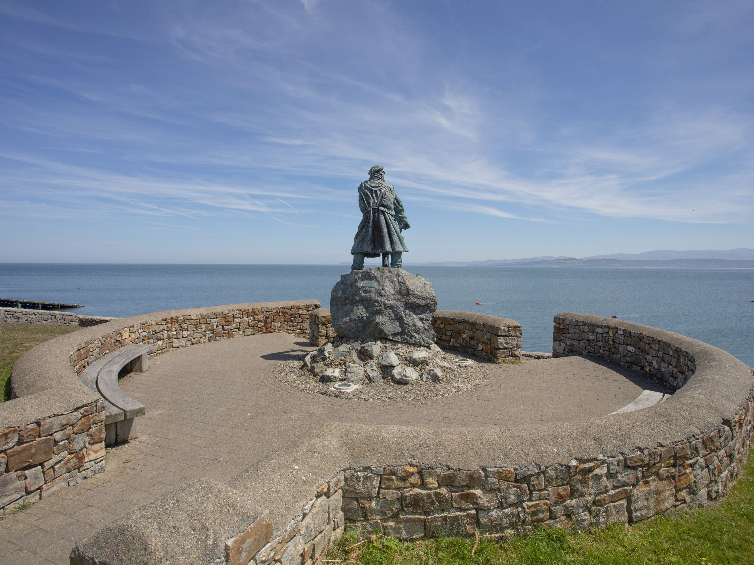 A statue on a stone pedestal surrounded by a circular stone wall with a view of the sea in the background.