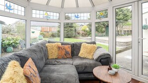 A conservatory with a sectional sofa, decorative pillows, and a small table with a plant. - Bryn Peris (Moelfre)