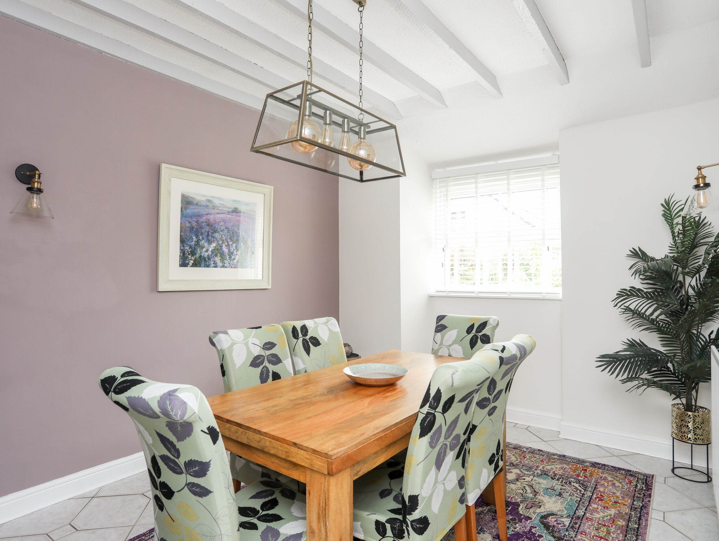 A dining area with a wooden table and chairs, a hanging light fixture, a framed painting, and a window with blinds.