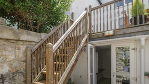 Eine Holztreppe führt zu einer Tür, hinter der eine steinerne Wand und ein Garten zu sehen sind.