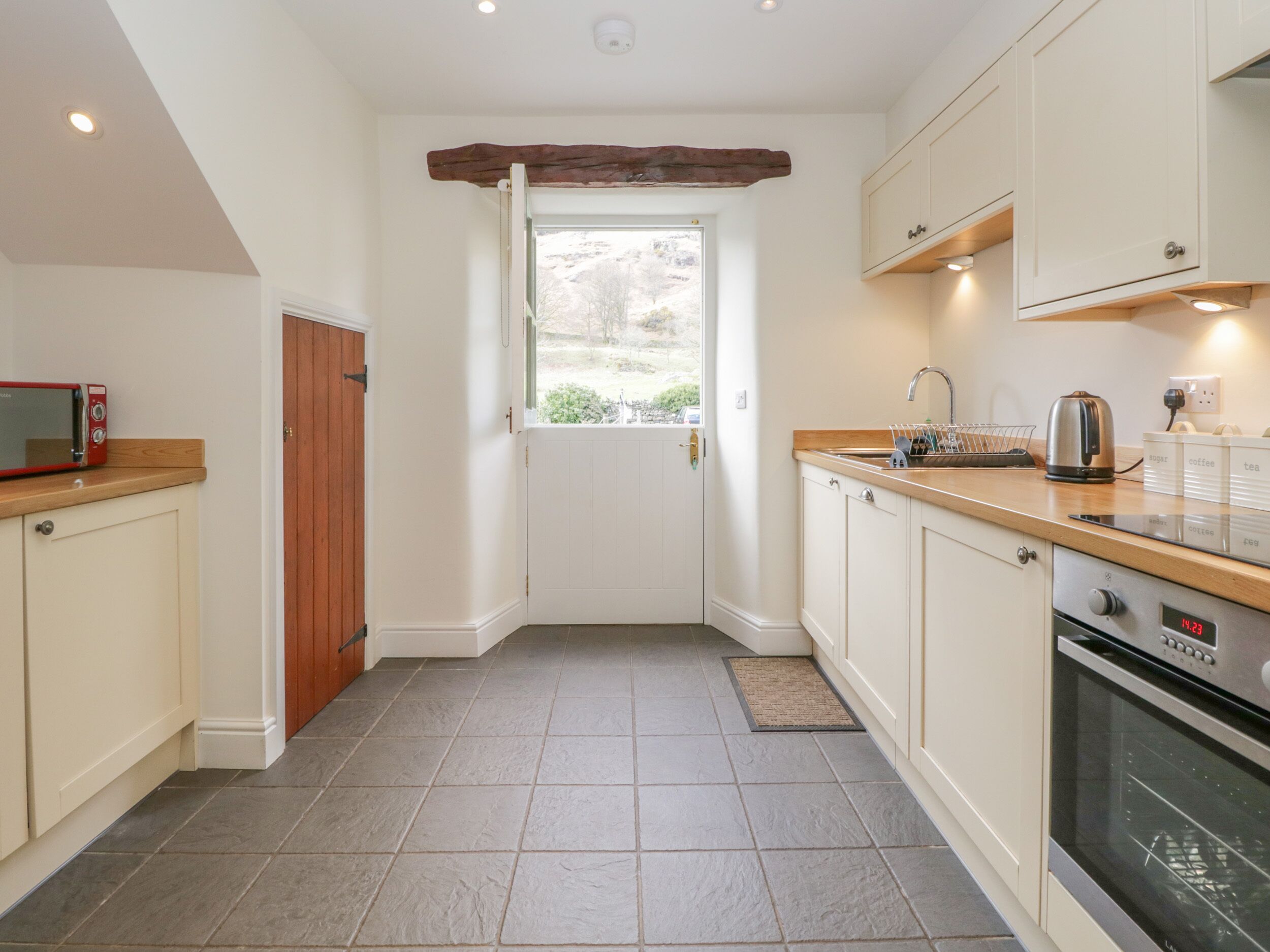 A compact kitchen with a microwave, kettle, and coffee maker on the counter, a wooden door, and a window with a view of greenery.