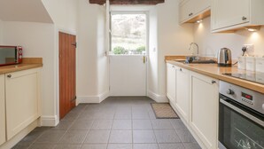 A compact kitchen with a microwave, kettle, and coffee maker on the counter, a wooden door, and a window with a view of greenery. - Holly Tree Cottage (Coniston)
