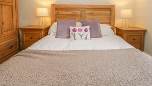 A neatly made bed with a beige blanket, two purple pillows, and a decorative pillow with a flower design. There are two wooden nightstands with lamps, a wooden wardrobe, and a wooden headboard. - Holly Tree Cottage (Coniston)