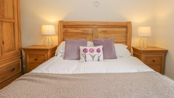 A neatly made bed with a beige blanket, two purple pillows, and a decorative pillow with a flower design. There are two wooden nightstands with lamps, a wooden wardrobe, and a wooden headboard.